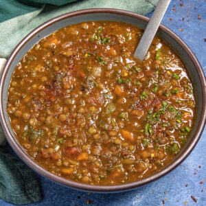A bowl of hearty Lentil Soup with visible vegetables and herbs, garnished with fresh parsley, sits on a blue surface beside a green cloth. A spoon rests in the flavorful soup.