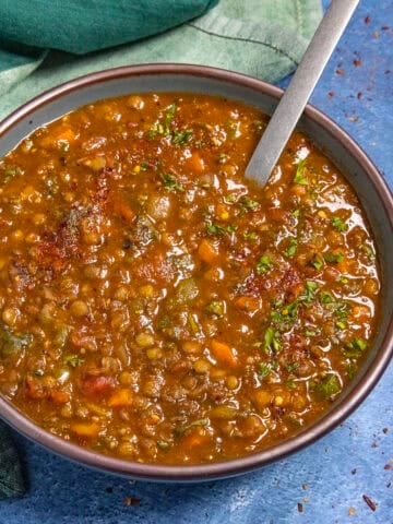 A bowl of hearty Lentil Soup with visible chopped vegetables and herbs sits on a blue surface, a spoon resting in the bowl and a green napkin beside it.
