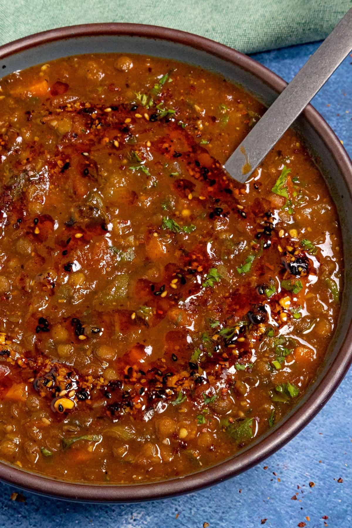 A close-up of Lentil Soup garnished with chopped herbs and a drizzle of chili oil, with a spoon resting inside the bowl on a blue surface.