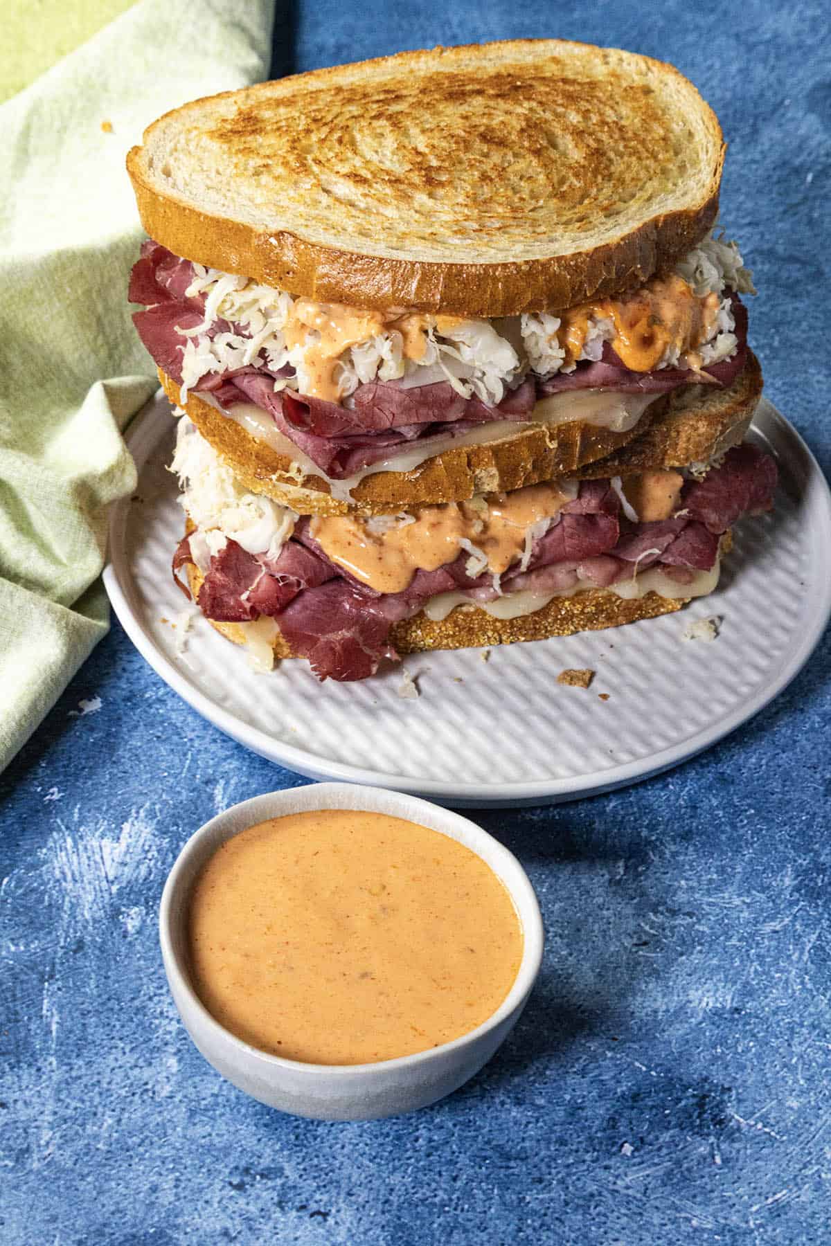 A plate with a stacked Reuben sandwich filled with corned beef, sauerkraut, Swiss cheese, and Russian Dressing sits beside a small bowl of creamy sauce on a blue textured surface.