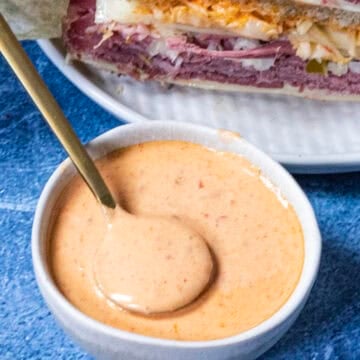 A small white bowl filled with creamy, pinkish-orange Russian Dressing and a spoon resting inside, placed on a blue surface next to a plate stacked with layered deli sandwiches.
