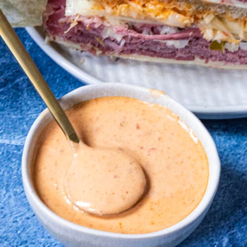 A small white bowl filled with creamy, pinkish-orange Russian Dressing and a spoon resting inside, placed on a blue surface next to a plate stacked with layered deli sandwiches.