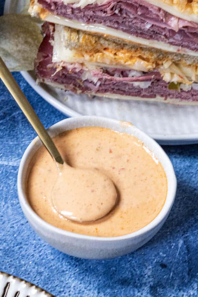 A small white bowl filled with creamy, pinkish-orange Russian Dressing and a spoon resting inside, placed on a blue surface next to a plate stacked with layered deli sandwiches.