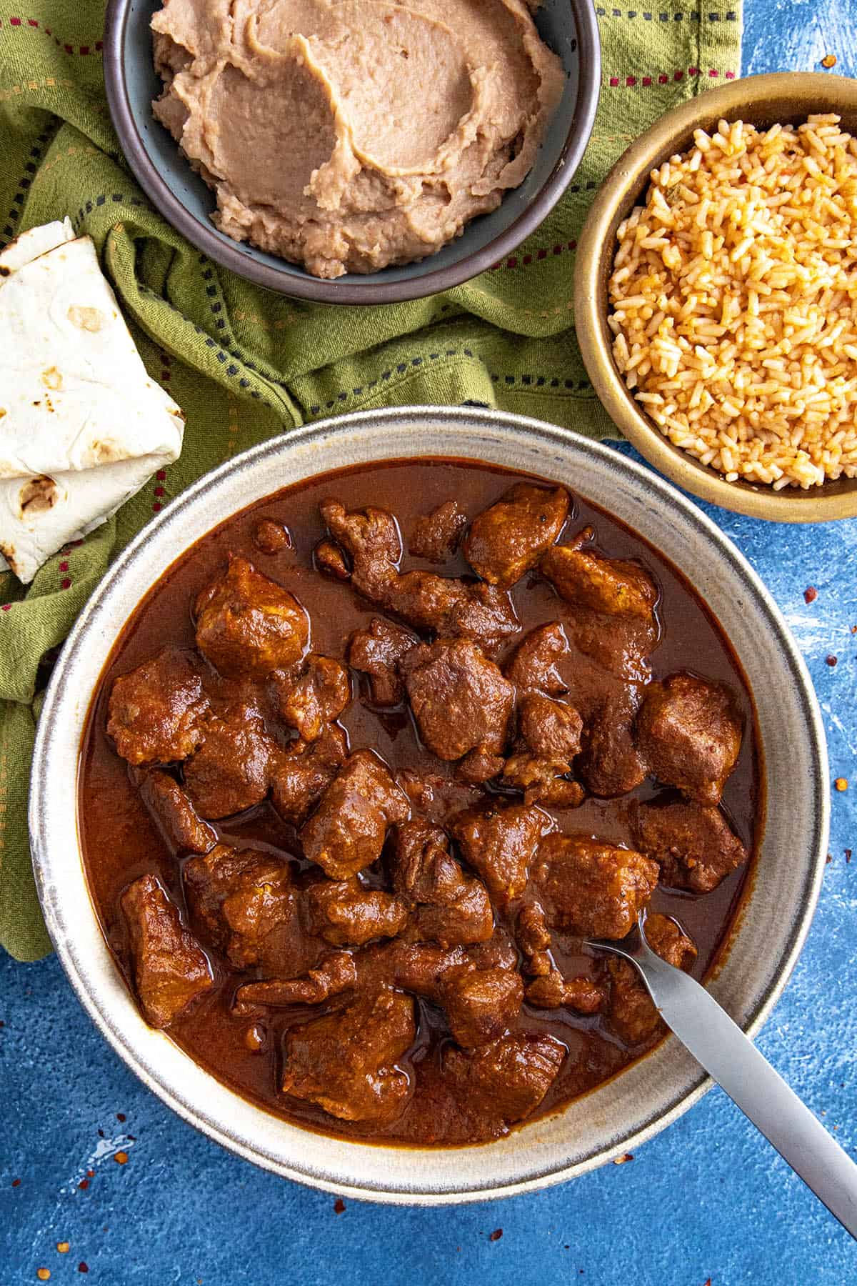 A saucy bowl of Mexican pork stew (asado de puerco) served with sides of Mexican rice, refried beans, and warmed tortillas