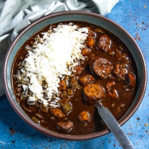 A bowl of dark, rich Cajun Chicken and Sausage Gumbo with sliced sausage, chunks of meat, and vegetables is served with a mound of white rice on top. A spoon rests in the bowl, and a light green napkin is nearby on a blue surface.