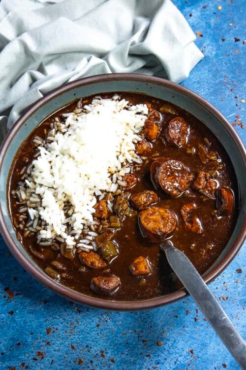 A bowl of Cajun Chicken and Sausage Gumbo, rich with dark broth, sausage slices, chicken, and vegetables, is topped with a mound of white rice. It sits on a blue surface beside a folded light gray cloth, with a spoon resting in the bowl.