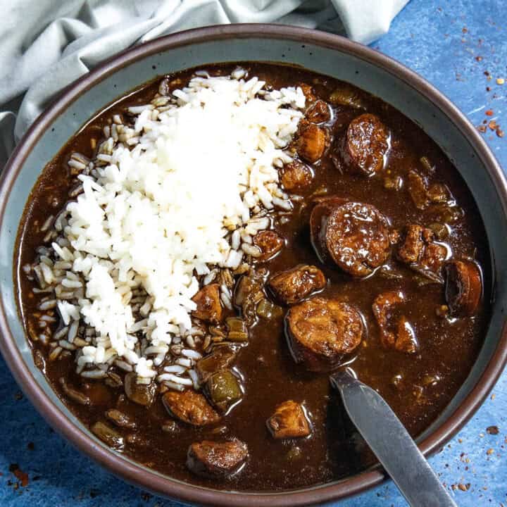 A bowl of Cajun Chicken and Sausage Gumbo, rich with dark broth, sausage slices, chicken, and vegetables, is topped with a mound of white rice. It sits on a blue surface beside a folded light gray cloth, with a spoon resting in the bowl.