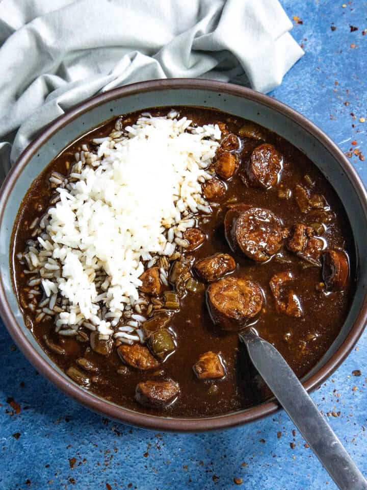 A bowl of Cajun Chicken and Sausage Gumbo, rich with dark broth, sausage slices, chicken, and vegetables, is topped with a mound of white rice. It sits on a blue surface beside a folded light gray cloth, with a spoon resting in the bowl.