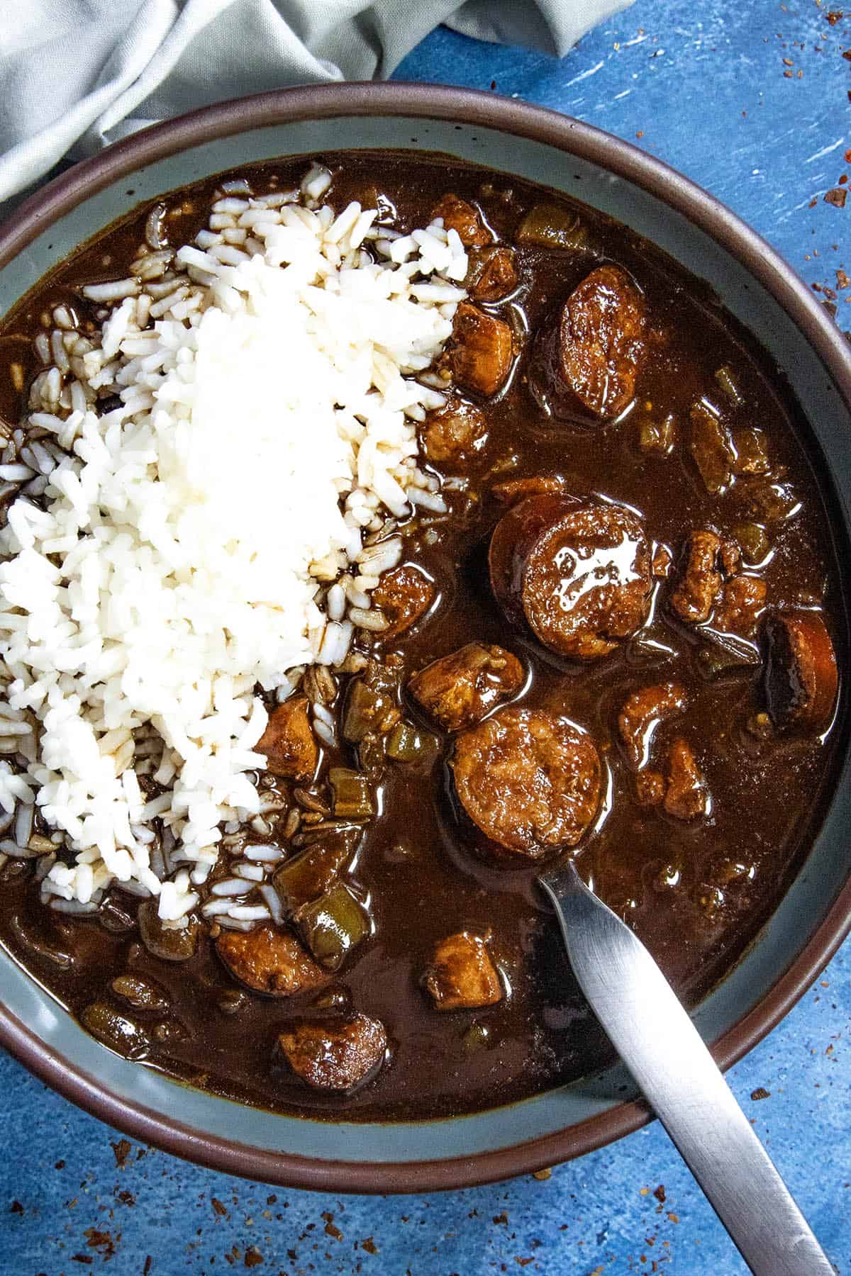 A bowl of dark, rich Cajun Chicken and Sausage Gumbo with sliced sausage, green peppers, and onions, served with a mound of white rice on top; a spoon rests in the bowl against a blue background.