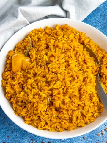 A white bowl loaded up with vibrant jollof rice, with a serving spoon, and a Scotch bonnet chili on top