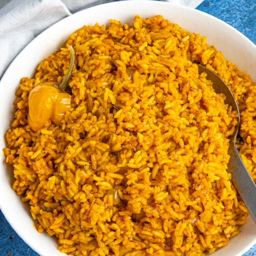 A white bowl loaded up with vibrant jollof rice, with a serving spoon, and a Scotch bonnet chili on top