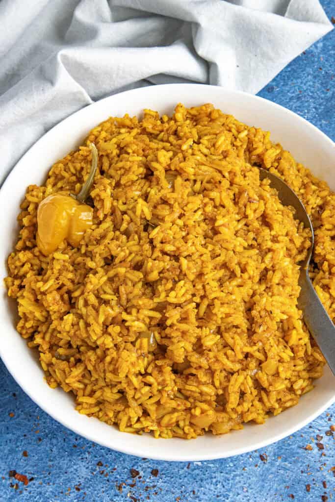 A white bowl loaded up with vibrant jollof rice, with a serving spoon, and a Scotch bonnet chili on top