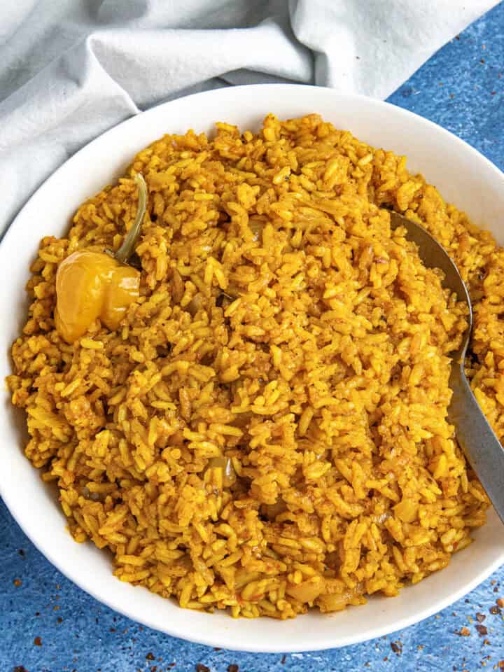 A white bowl loaded up with vibrant jollof rice, with a serving spoon, and a Scotch bonnet chili on top