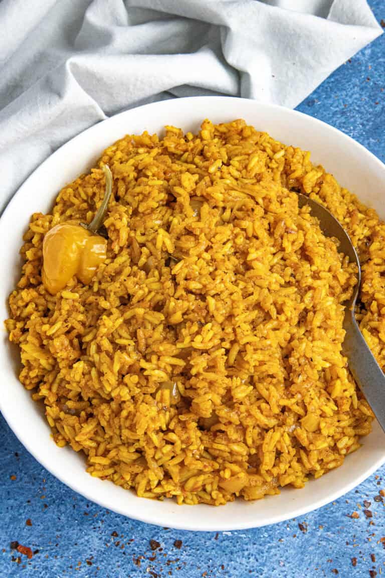 A white bowl loaded up with vibrant jollof rice, with a serving spoon, and a Scotch bonnet chili on top