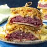 Two halves of a classic Reuben Sandwich stacked on a plate, showing layers of corned beef, Swiss cheese, sauerkraut, and dressing on toasted rye bread, served with crunchy potato chips on the side.