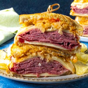 Two halves of a classic Reuben Sandwich stacked on a plate, showing layers of corned beef, Swiss cheese, sauerkraut, and dressing on toasted rye bread, served with crunchy potato chips on the side.