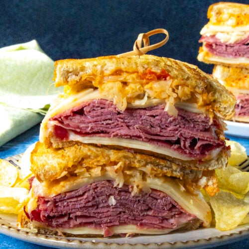 A close-up of a classic Reuben Sandwich cut in half, stacked on a plate with layers of corned beef, Swiss cheese, sauerkraut, and dressing on rye bread, served with crispy potato chips on the side.