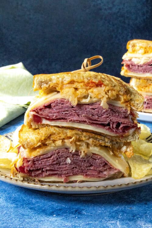 A close-up of a classic Reuben Sandwich cut in half, stacked on a plate with layers of corned beef, Swiss cheese, sauerkraut, and dressing on rye bread, served with crispy potato chips on the side.