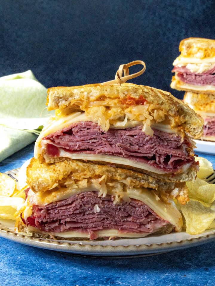 A close-up of a classic Reuben Sandwich cut in half, stacked on a plate with layers of corned beef, Swiss cheese, sauerkraut, and dressing on rye bread, served with crispy potato chips on the side.