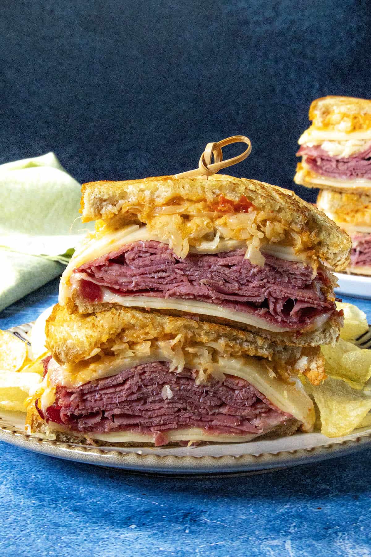 A close-up of a classic Reuben Sandwich cut in half, stacked on a plate with layers of corned beef, Swiss cheese, sauerkraut, and dressing on rye bread, served with crispy potato chips on the side.