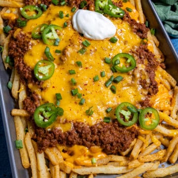 A sheet pan of Chili Cheese Fries piled high with French fries, chili, melted cheddar, jalape&ntilde;os, green onions, and a dollop of sour cream. A green cloth napkin sits in the corner.