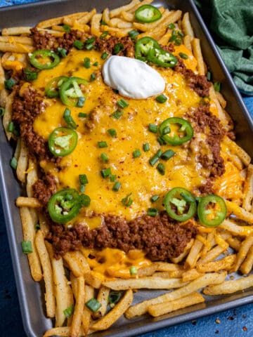 A sheet pan of Chili Cheese Fries piled high with French fries, chili, melted cheddar, jalapeños, green onions, and a dollop of sour cream. A green cloth napkin sits in the corner.