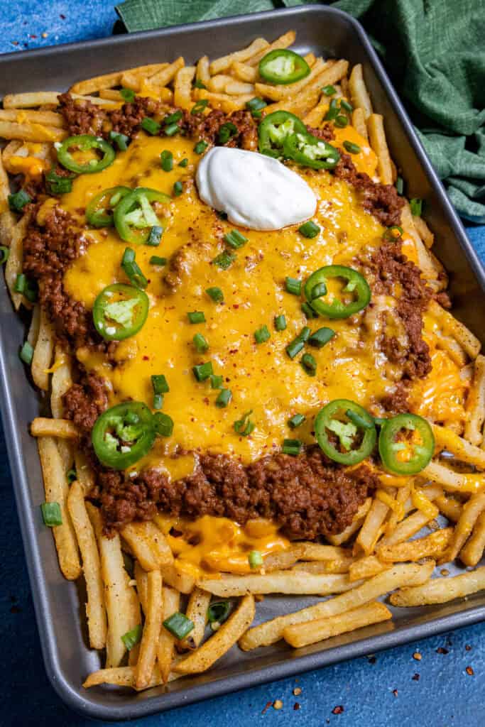 A sheet pan of Chili Cheese Fries piled high with French fries, chili, melted cheddar, jalapeños, green onions, and a dollop of sour cream. A green cloth napkin sits in the corner.