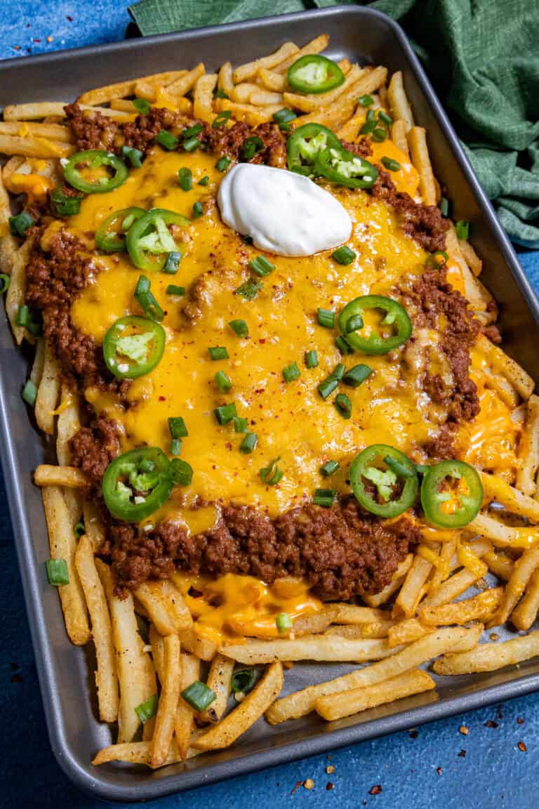 A sheet pan of Chili Cheese Fries piled high with French fries, chili, melted cheddar, jalape&ntilde;os, green onions, and a dollop of sour cream. A green cloth napkin sits in the corner.