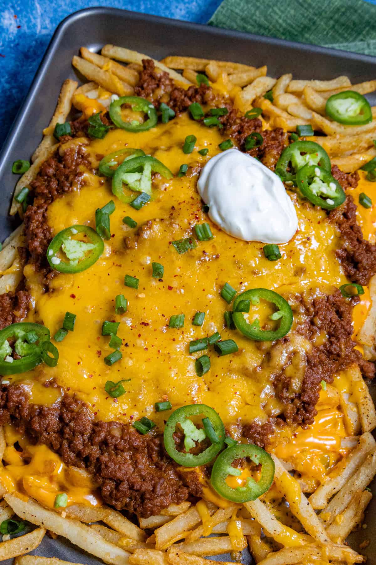 A tray of chili cheese fries loaded with melted cheddar, savory chili, sliced jalapeños, chopped green onions, and a dollop of sour cream.
