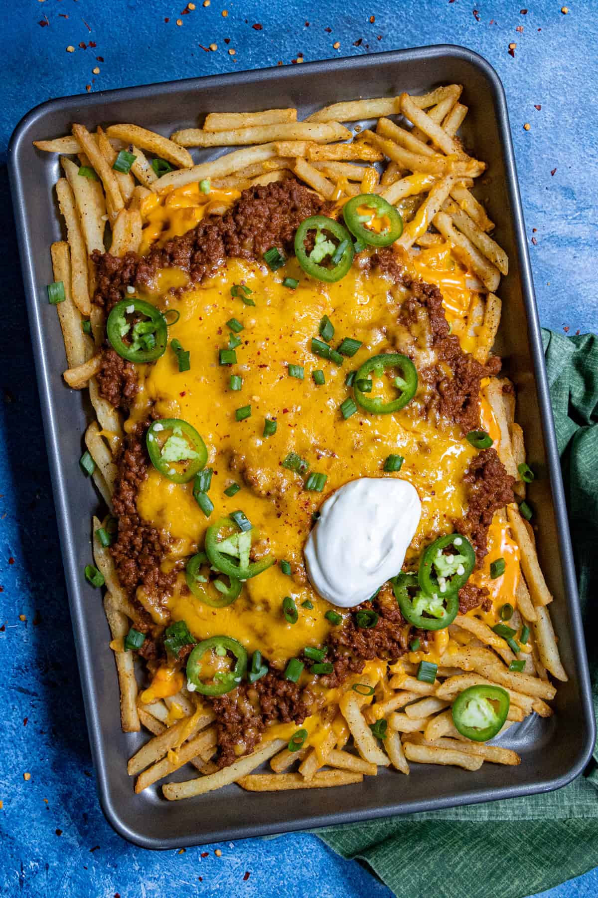 A large baking tray filled with Chili Cheese Fries, with meaty chili and lots of melty cheese, topped with sour cream, sliced chilies, and green onion