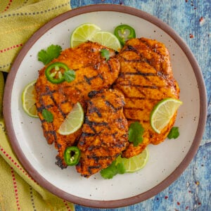 Three grilled Chili Lime Chicken breasts with grill marks are served on a white plate, garnished with lime slices, cilantro, and jalape&ntilde;o slices.