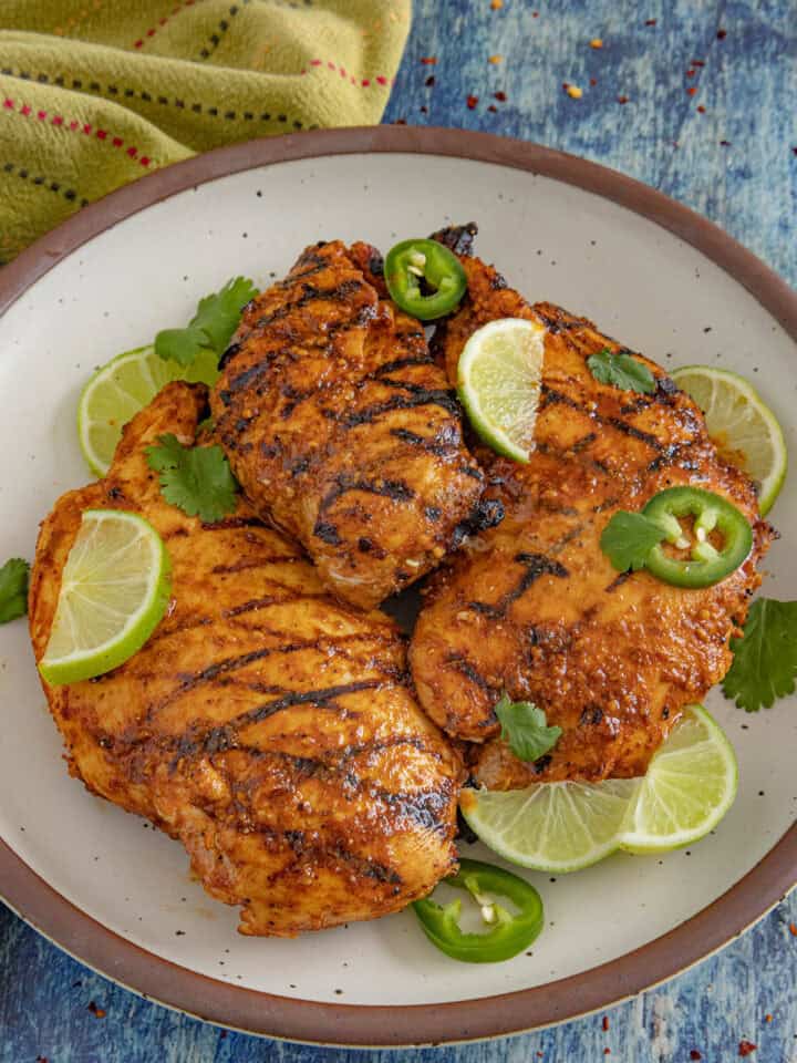 3 chili lime chicken breasts with char grill marks on a plate, garnished with lime, cilantro, and sliced jalapeno