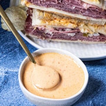 A white bowl of creamy Russian Dressing with a spoon. Behind it, two stacked Reuben sandwiches filled with sliced meat, cheese, and cabbage sit on a white plate.