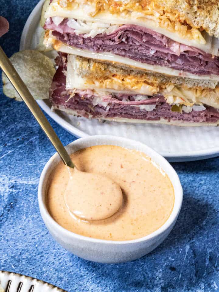 A white bowl of creamy Russian Dressing with a spoon. Behind it, two stacked Reuben sandwiches filled with sliced meat, cheese, and cabbage sit on a white plate.