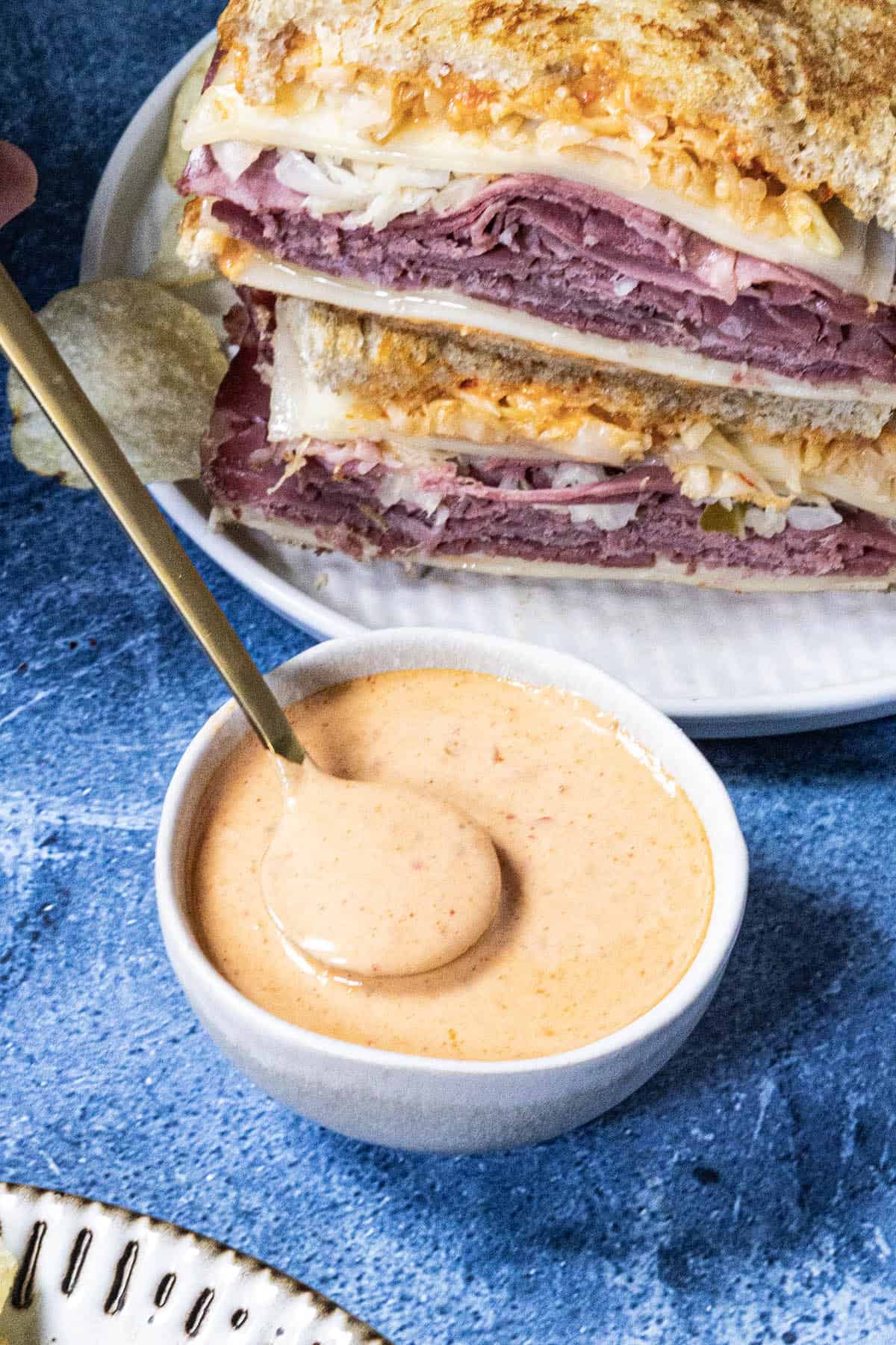 A white bowl of creamy Russian Dressing with a spoon. Behind it, two stacked Reuben sandwiches filled with sliced meat, cheese, and cabbage sit on a white plate.