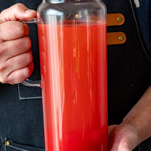 Mike in a dark apron holding a clear glass pitcher filled with bright red Agua Fresca.