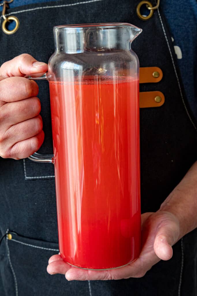 Mike in a dark apron holding a clear glass pitcher filled with bright red Agua Fresca.