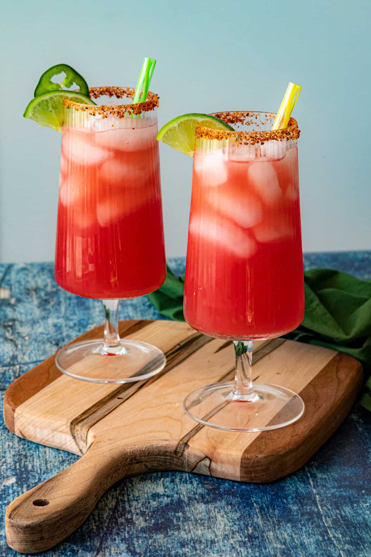Two tall glasses of homemade Agua Fresca, filled with a red iced drink, garnished with lime slices, chili powder rims, a jalape&ntilde;o slice, and colorful straws, set on a wooden board against a blue background.