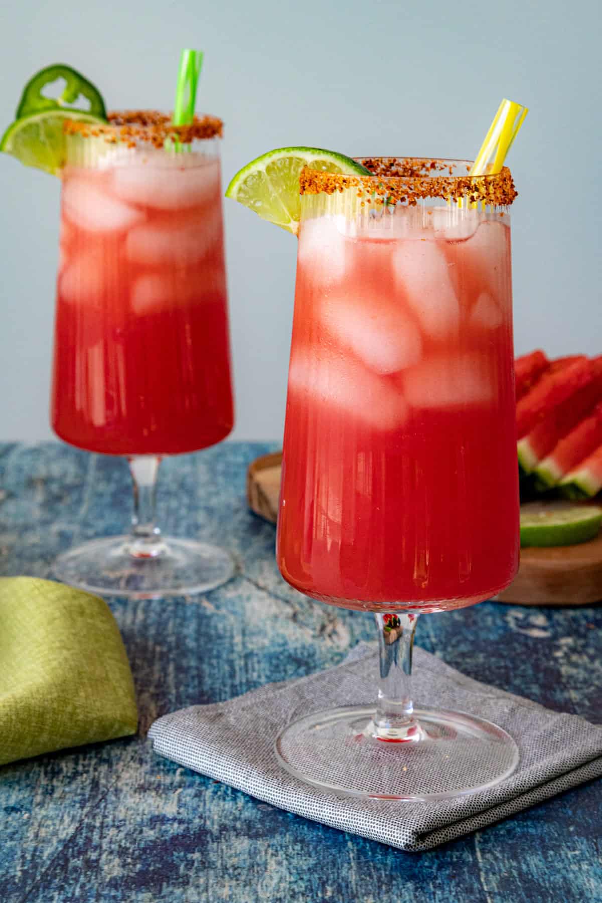 Two tall glasses of Watermelon Agua Fresca with green and yellow straws, garnished with lime wedges and seasoning on the rims, sit on a blue surface with a cloth napkin. Slices of watermelon and lime are in the background.
