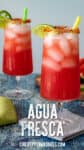 Two tall glasses of pink-red Agua Fresca with lime wedges, tajin-rimmed edges, and green and yellow straws rest on a wooden board against a blue background.