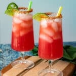 Two tall glasses of pink-red Agua Fresca with lime wedges, tajin-rimmed edges, and green and yellow straws rest on a wooden board against a blue background.