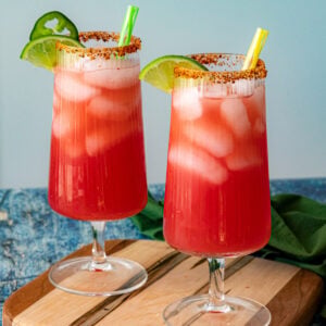 Two tall glasses of pink-red Agua Fresca with lime wedges, tajin-rimmed edges, and green and yellow straws rest on a wooden board against a blue background.