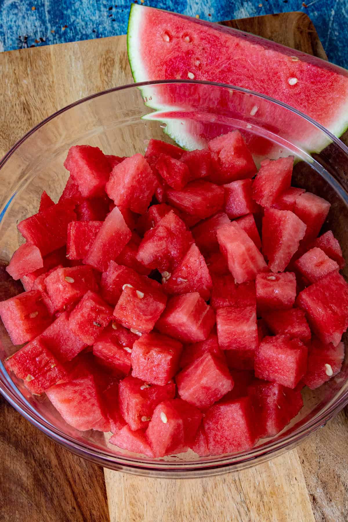 A glass bowl filled with cubed watermelon, perfect for making Agua Fresca, sits on a wooden surface. A wedge of watermelon with rind is placed beside the bowl.