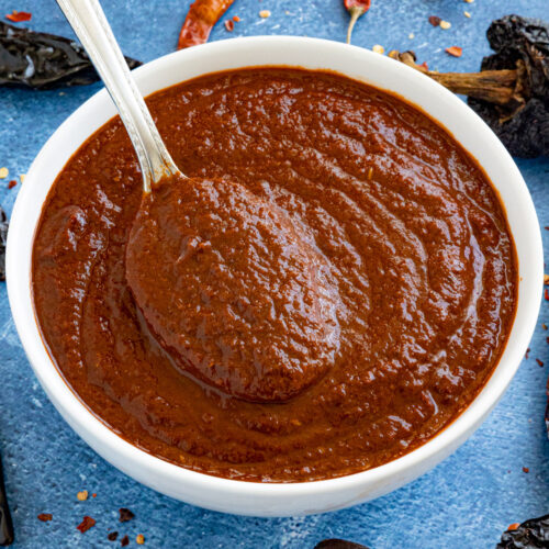 A white bowl filled with thick, dark red Birria sauce, with a spoon resting inside. Dried chili peppers and red pepper flakes are scattered on a blue surface around the bowl.