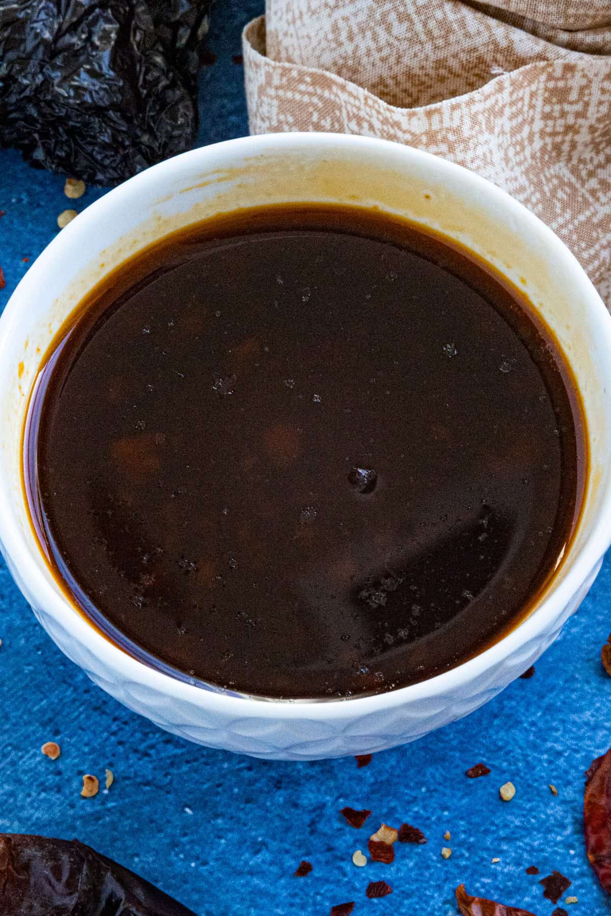 A white bowl filled with dark brown birria sauce sits on a blue surface, with a beige cloth and dried chili peppers nearby.