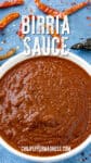 A bowl filled with rich, red Birria Sauce sits on a blue surface, surrounded by dried chili peppers. Large text above the bowl reads Birria Sauce and the website chilipeppermadness.com appears at the bottom.