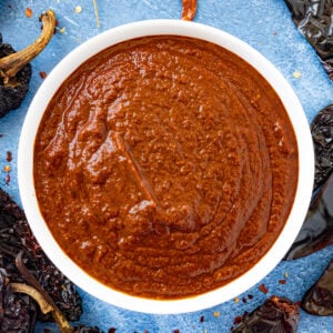 A white bowl filled with thick, rich red Birria Sauce sits on a blue surface, surrounded by dried chili peppers.
