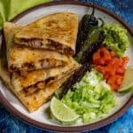 A plate with Chicken Quesadillas, grilled peppers, guacamole, chopped tomatoes, shredded lettuce, and lime wedges is served on a speckled plate against a blue background.