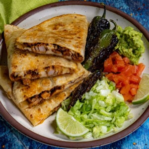 A plate with Chicken Quesadillas, grilled peppers, guacamole, chopped tomatoes, shredded lettuce, and lime wedges is served on a speckled plate against a blue background.