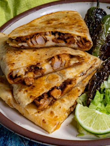 A plate with three stacked homemade Chicken Quesadillas, a side of grilled peppers, shredded lettuce, and lime wedges on a brown-rimmed dish against a blue background.